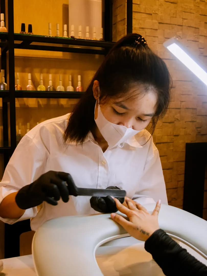 A technician in a white shirt, mask, and black gloves filing a client’s nails over a pale basin.