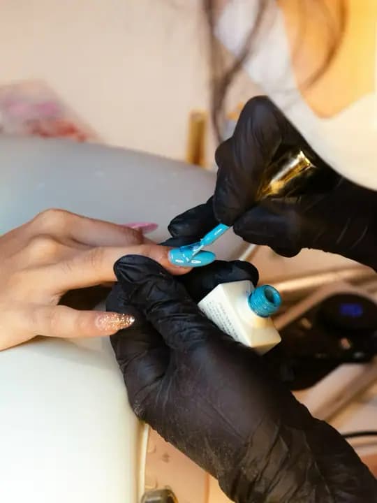 Gloved hands applying turquoise gel polish to a client’s nail.