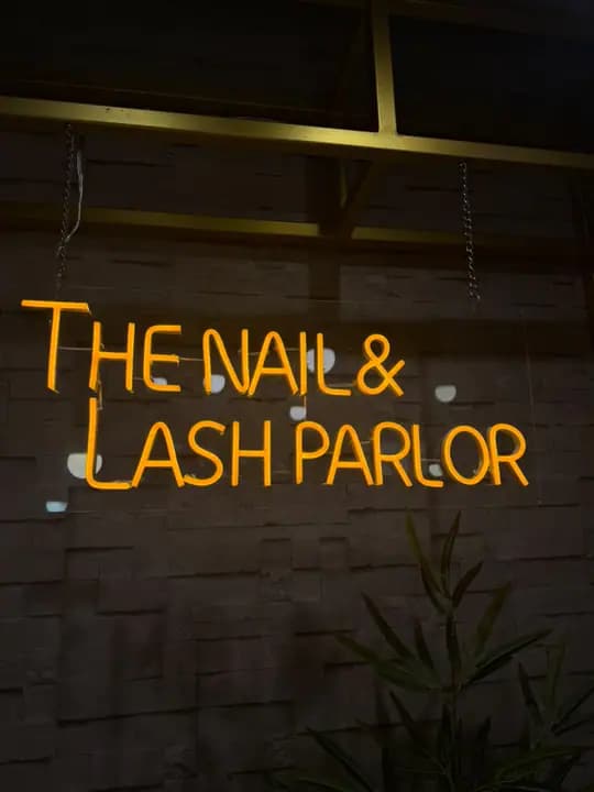 Neon sign on a stone wall that reads The Nail and Lash Parlor, with a plant below it.
